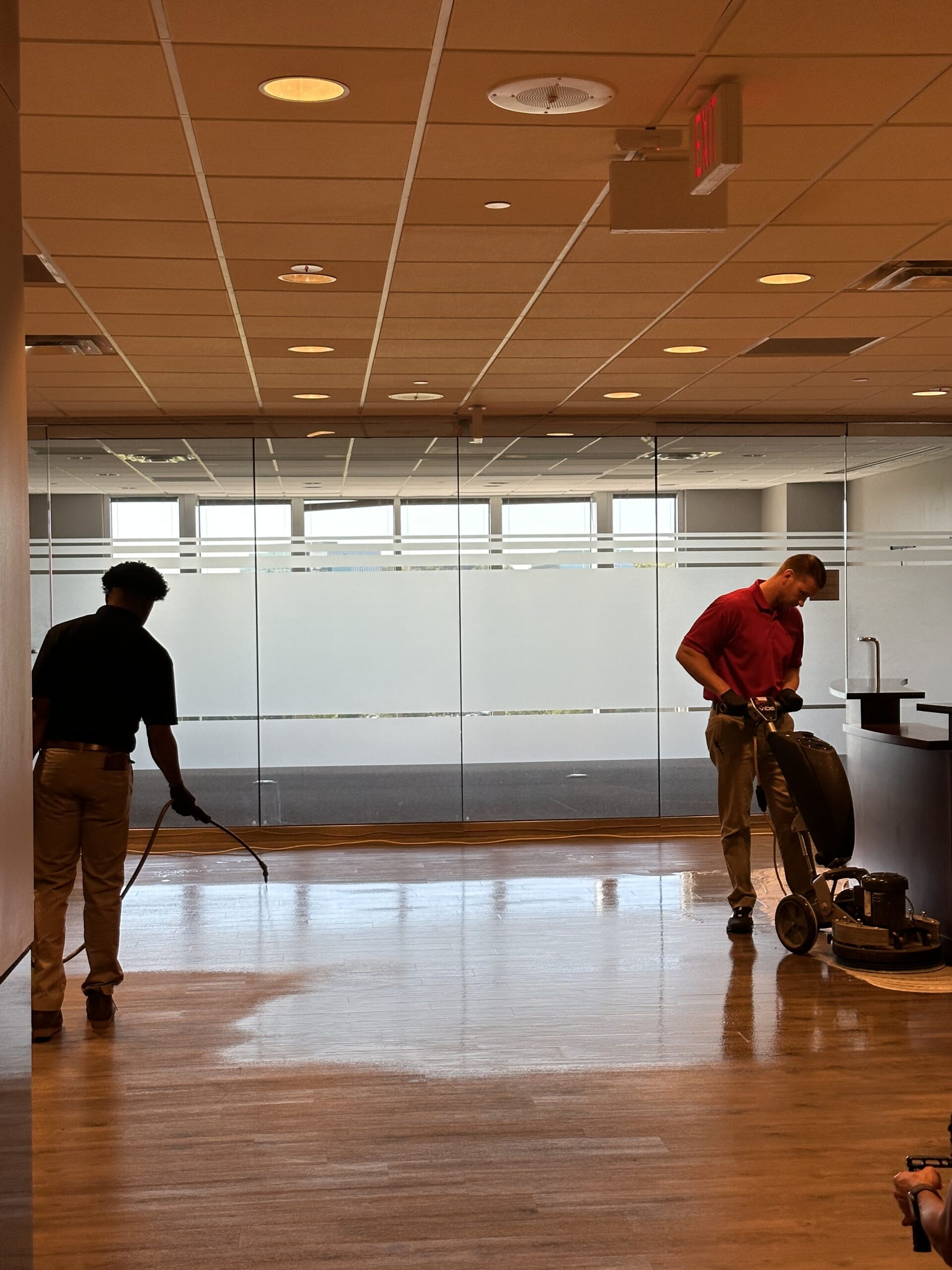 Two men cleaning a commercial office's floor with one spraying the floor with soap and the other vacuuming. DPM Surface Care